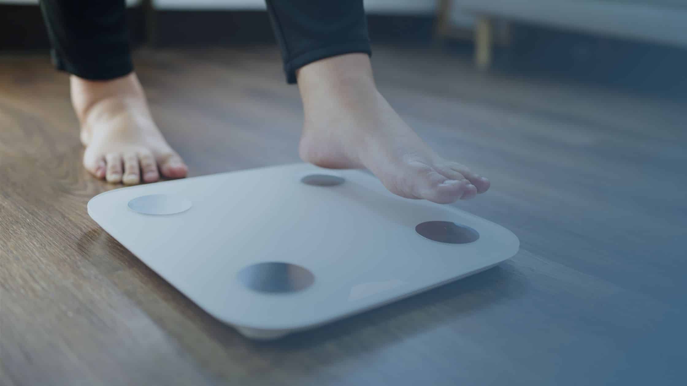 Pessoa descalça prestes a subir em uma balança digital, sobre um piso de madeira, com foco nos pés e na balança.
